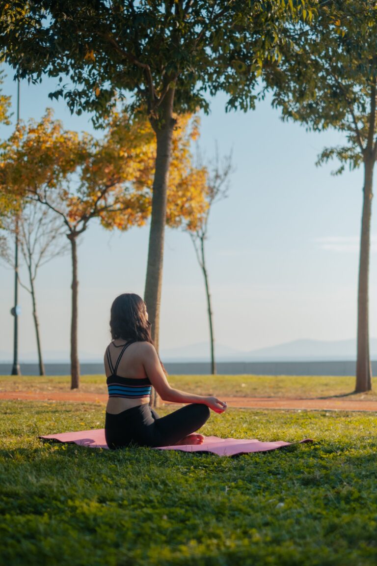 A woman meditates outdoors on a yoga mat in a tranquil park setting, surrounded by nature.